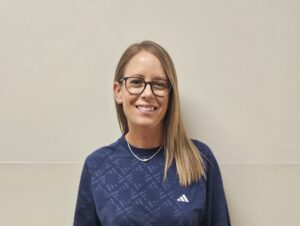 Woman with glasses and long blond hair, wearing a navy patterned top and light gray cargo pants, smiling at the camera against a beige wall.