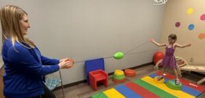 Young girl in a purple leotard balances on colorful stepping stones, tied to an adult with a resistance band in a therapy room.