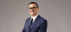 Professional headshot: smiling man in a navy suit, white shirt, and striped tie, wearing glasses against a neutral gray backdrop.
