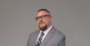 Head-and-shoulders portrait of a bearded man with short hair, wearing black glasses and a gray suit against a neutral backdrop.