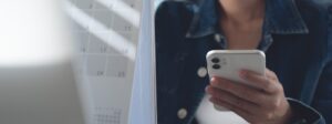 Person in a denim jacket using a smartphone at a desk with a large calendar in the blurred background, conveying a workspace moment. (informative)